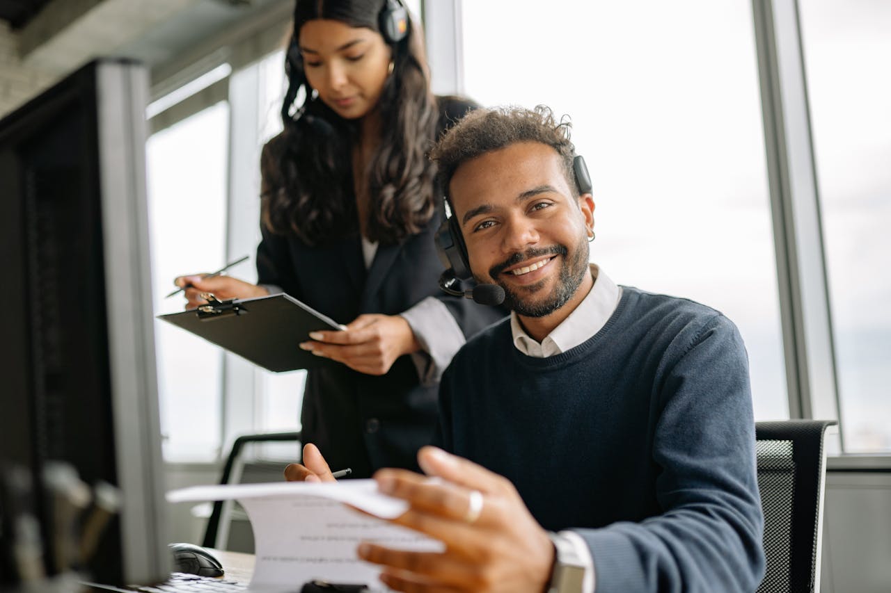 Friendly call center agent wearing a headset, talking and smiling at his desk with a colleague.